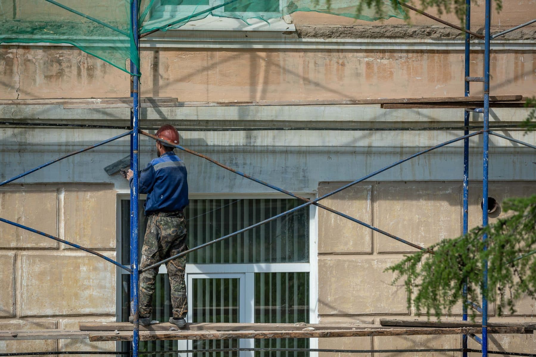 A worker in a hard hat on scaffolding, working on the stone facade of a building.
