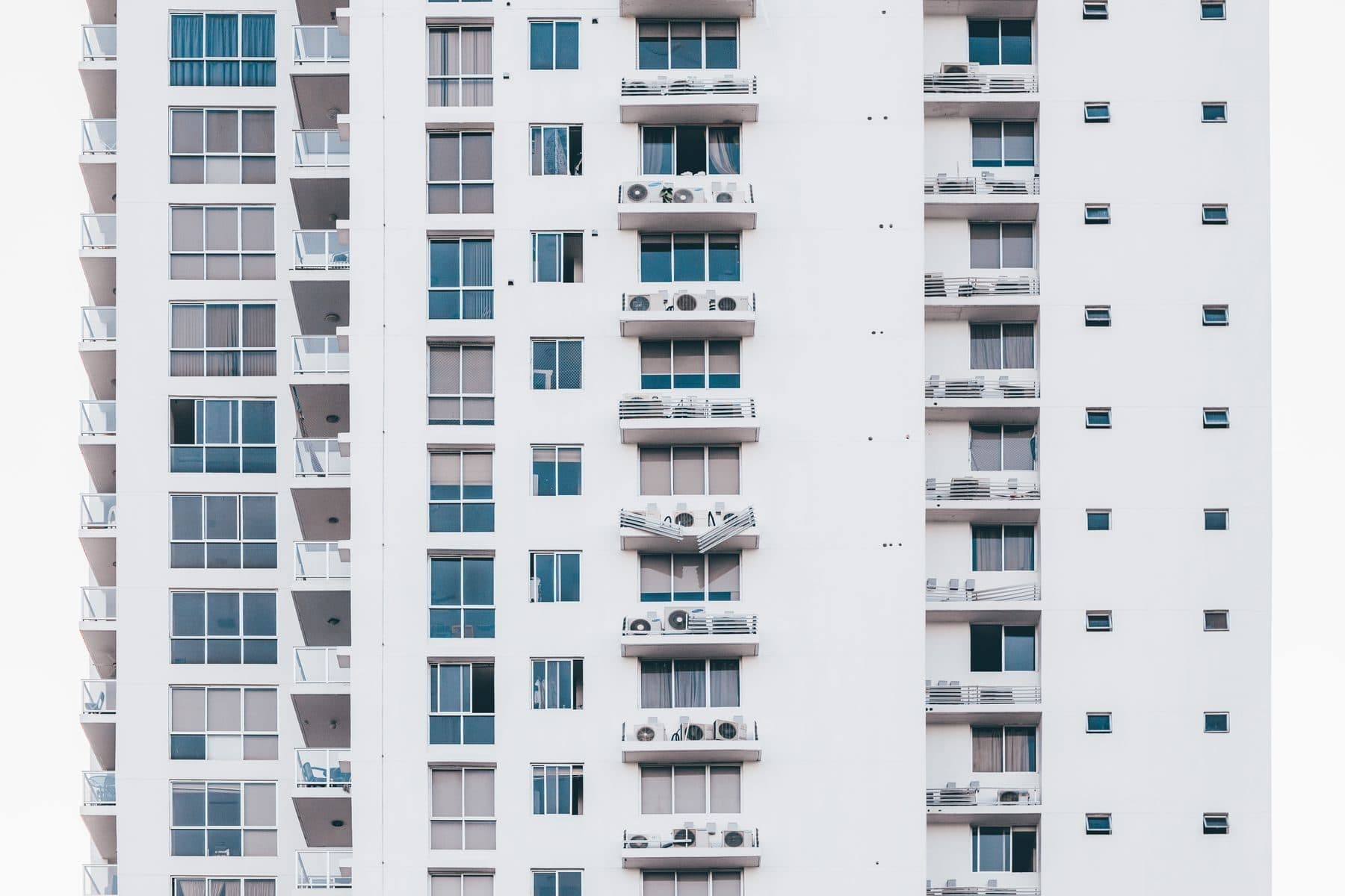 A white residential tower with stacked balconies and rows of wall air conditioners.
