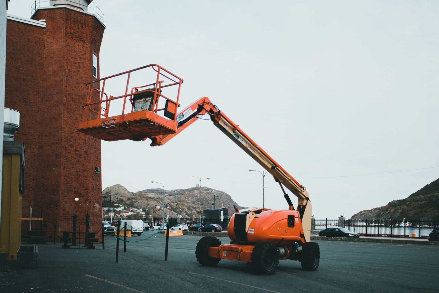 An orange boom lift with its basket raised beside a red brick building.