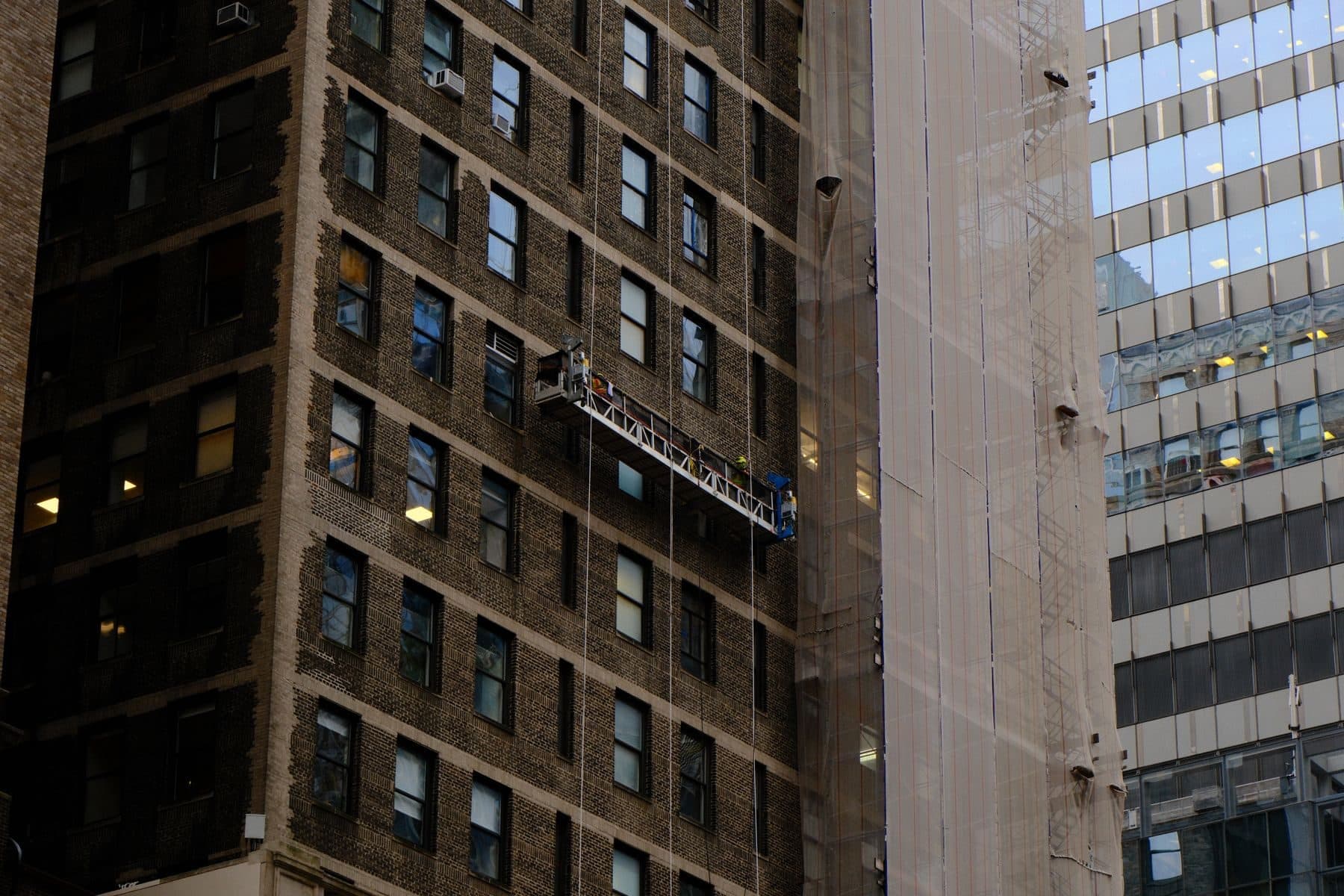 Workers on a suspended platform hanging against the face of a tall brick building.