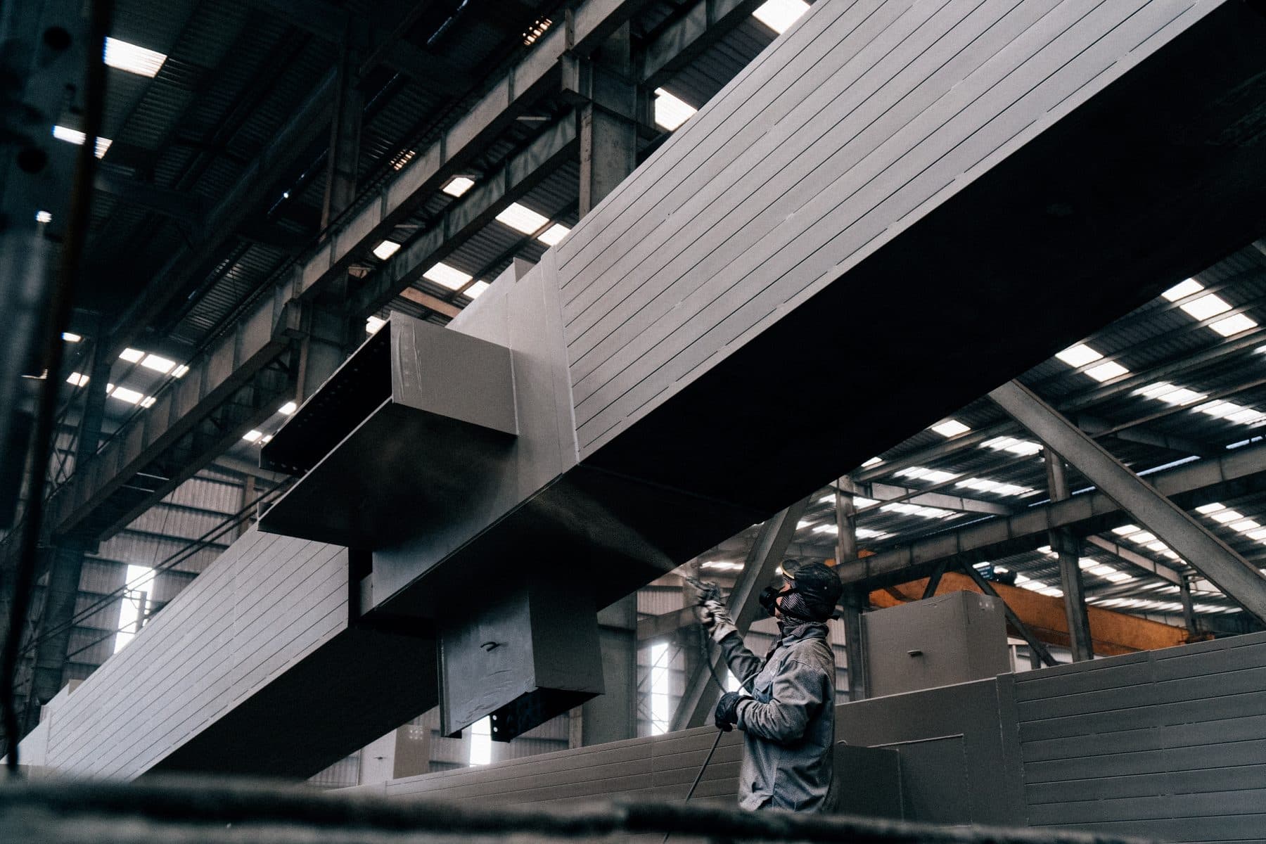A worker in a respirator spraying coating onto large steel beams inside a warehouse.